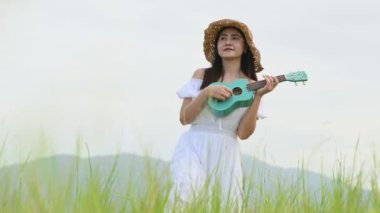 Beyaz elbiseli Asyalı mutlu kadın yeşil çayır tarlasında gitar çalıyor ve yaz mevsiminde dağcılık yapıyor. Müzisyen güzel kadın müzik aleti ile rahatla. Kırsal kesimde yaşam tarzı bağbozumu