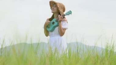 Beyaz elbiseli Asyalı mutlu kadın yeşil çayır tarlasında gitar çalıyor ve yaz mevsiminde dağcılık yapıyor. Müzisyen güzel kadın müzik aleti ile rahatla. Kırsal kesimde yaşam tarzı bağbozumu