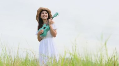 Beyaz elbiseli Asyalı mutlu kadın yeşil çayır tarlasında gitar çalıyor ve yaz mevsiminde dağcılık yapıyor. Müzisyen güzel kadın müzik aleti ile rahatla. Kırsal kesimde yaşam tarzı bağbozumu