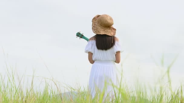 身穿白衣的亚洲快乐女人在绿茵的草地上弹奏着四弦琴 背景是隆重的 音乐家的美女用乐器放松 农村的终身制 — 图库视频影像