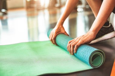 Spor salonunun arka planında yoga şiltesini katlayan sportif bir kadın. Yoga dersinden sonra paspas yuvarlama egzersizi. Spor ve spor eğitimi konsepti. Ellerini halıya koy.