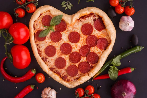 Pizza heart shaped with pepperoni with ingredients on black background. Concept of romantic love for Valentines Day . Love food