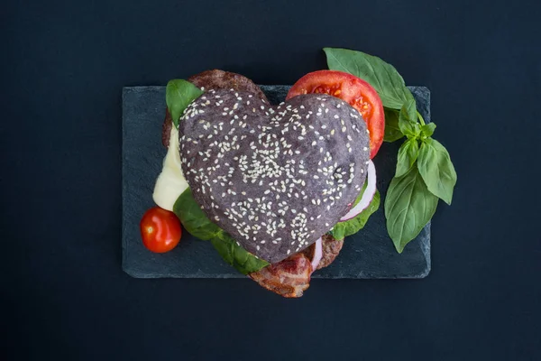 Heart shaped black hamburger on stone plate, love burger fast food concept, Valentines day surprise dinner, wooden background, top view flat lay