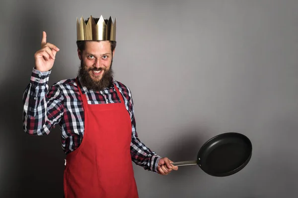King of cooking with finger up, funny bearded man in paper crown and cook apron with empty frying pan is ready to tech a lesson