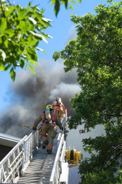 MONTREAL, CANADA - 18 Haziran 2020: İtfaiyeciler Montreal 'deki bir konut binasındaki yangını söndürmek için gökyüzüne doğru tırmanıyorlar. Dikiz manzarası merdiveni çevreleyen yaprakların arasından yukarıya, mavi gökyüzüne doğru siyah duman bulutlarına bakıyor.