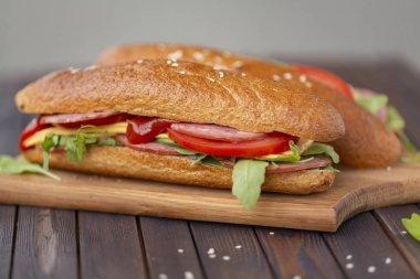 İki tane baget sandviç salam, peynir, roka salatası, domates ve soğan bir kesme tahtası üzerinde. Koyu ahşap arka plan üzerinde sandviç. Lezzetli Aperatif.