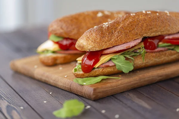 Two baguette sandwiches with salami, cheese, arugula salad, tomatoes and onion on a cutting board. Sandwiches on dark wooden background. Tasty snack.