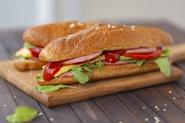 Two baguette sandwiches with salami, cheese, arugula salad, tomatoes and onion on a cutting board. Sandwiches on dark wooden background. Tasty snack.