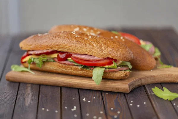 Two baguette sandwiches with salami, cheese, arugula salad, tomatoes and onion on a cutting board. Sandwiches on dark wooden background. Tasty snack.