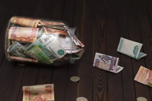 Glass jar full of money tipped over on its side spilling money on wooden background.