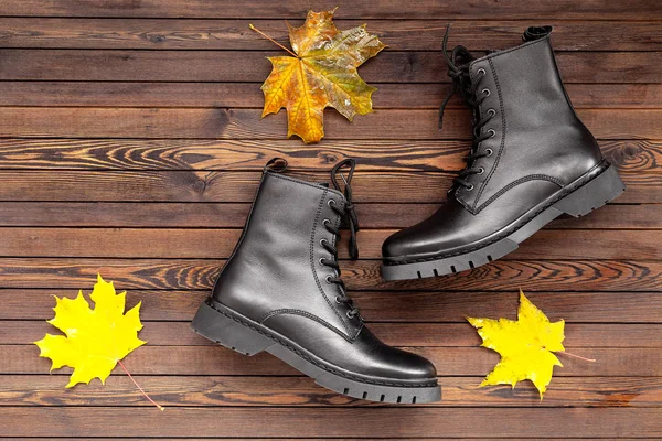 Women's autumn shoes. Fashionable Black rough boots for walks, on a dark wooden background with autumn leaves. Flat lay. Autumn Fashion Concept.