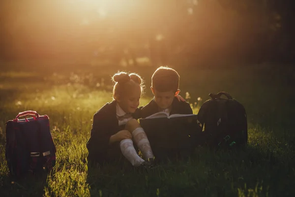 Małe dzieci w szkole czytać książkę, ubrany w mundurze szkolnym z torbą, siedząc na trawie po szkole, na zewnątrz, o zachodzie słońca , — Zdjęcie stockowe