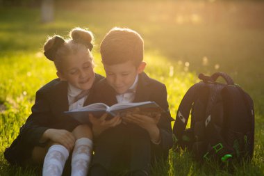 küçük okul çocukları, küçük çocuk ve küçük kız bir kitap okuma, okul çantası ile okul üniforması giymiş, okuldan sonra çim üzerinde oturan, açık havada, gün batımında