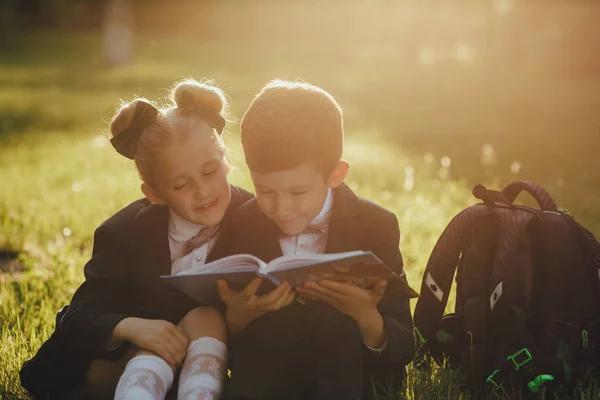 little school children, little boy and little girl reading a book, dressed in school uniform with schoolbag, sitting on grass after school, outdoor, at sunset