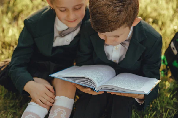 close up, little school children, little boy and little girl reading a book, dressed in school uniform with schoolbag, sitting on grass after school, outdoor, at sunset
