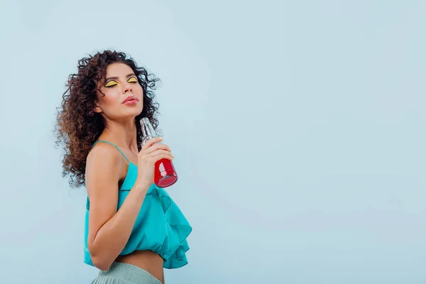 fashionable lady with curly hair, smiles has bottle with drink in hand, dressed in blue shirt, eyes closed, isolated on blue background, copy space