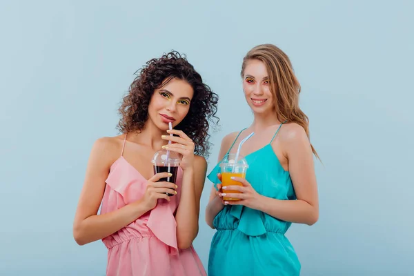 Две счастливые женщины с вьющимися волосами улыбаются и держать пластиковую чашку с соком в розовой и синей рубашке, положительные эмоции лица, изолированные на синем фоне, копировать пространство — стоковое фото