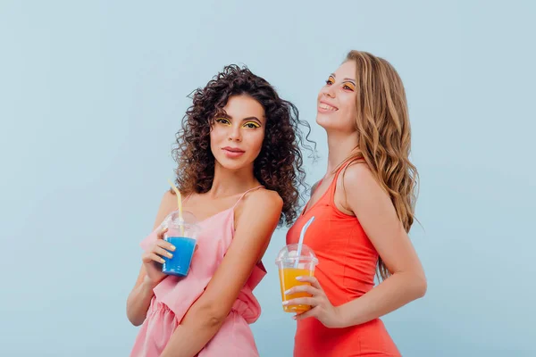 Модные две девушки с вьющимися волосами, держащие в руке пластиковую чашку с соком, одетые в розовую и красную футболки позитивные эмоции лица, изолированные на синем фоне, копировальное пространство — стоковое фото