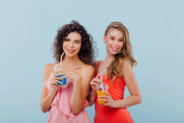 Модные две веселые девушки с вьющимися волосами, держать пластиковую чашку с соком, смотреть на камеру позитивные эмоции лица, изолированные на синем фоне, копировать пространство — стоковое фото