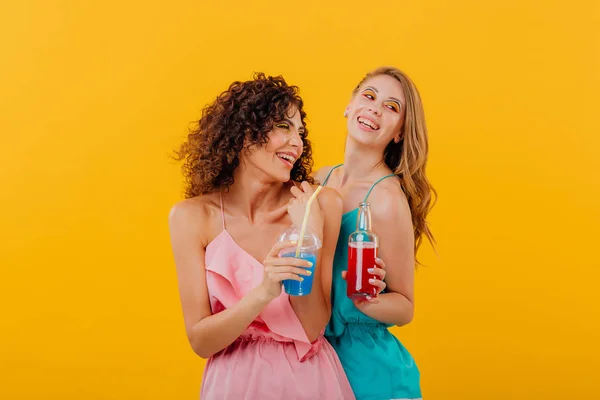 Dos chicas guapas con pelo rizado, divirtiéndose con la botella de bebida en la mano y taza de plástico con jugo, emociones faciales positivas, aisladas sobre fondo amarillo, espacio para copiar — Foto de Stock