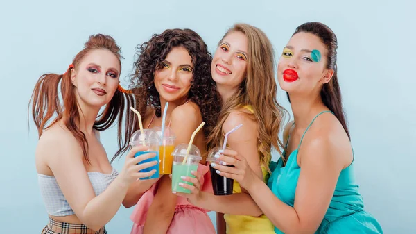 Четыре молодые девушки волосы и макияж необычно, держит пластиковые чашки сока, посмотрите на камеру, изолированный синий фон, положительные эмоции лица, копировать пространство — стоковое фото