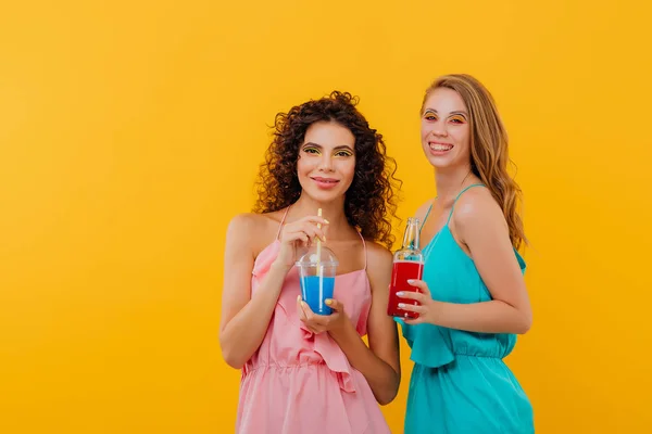 Dos hermosas chicas con pelo rizado bebida botella en la mano y taza de plástico con jugo, mira a la cámara, emociones faciales positivas, aislado en el fondo amarillo, espacio de copia — Foto de Stock