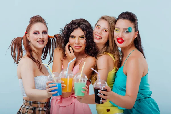 Четыре счастливые молодые девушки с необычной прической и макияжем, держа пластиковые стаканчики сока, глядя на камеру изолированный синий фон, положительные эмоции лица, копировать пространство — стоковое фото