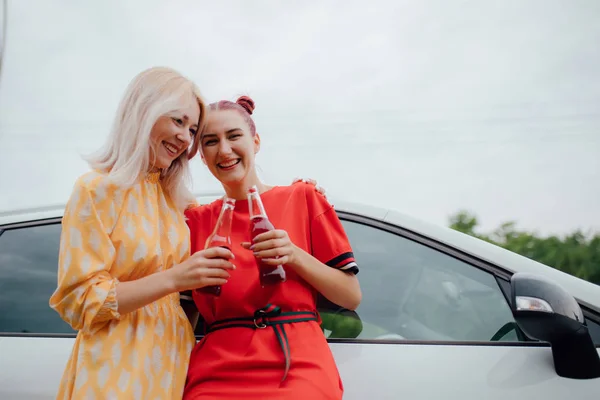 Dvě přítelkyně jsou veselé, baví se v červených a žlutých šatech, má v ruce láhev s nápoji. vedle vozu, venkovní — Stock fotografie