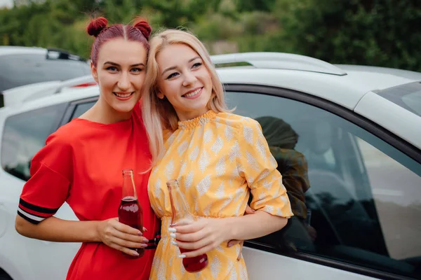 Duas belas mulheres jovens, se divertindo em vestidos vermelhos e amarelos, tem a garrafa de bebida em sua mão. próximo ao carro, ao ar livre — Fotografia de Stock