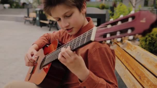 Närbild, barn med gitarr sitter på en stol i parken, klädd i en skjorta, — Stockvideo