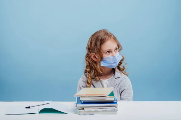 schoolgirl with medical mask on her face, has books on the table