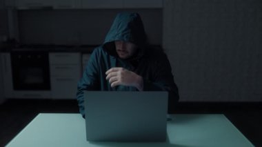 Karanlık bir odada kapüşonlu kazak giyen, evde dizüstü bilgisayar üzerinde çalışan bir bilgisayar korsanı. Modern siber güvenlik tehditlerini anlatan tehlikeli ve gizemli bir sahne..