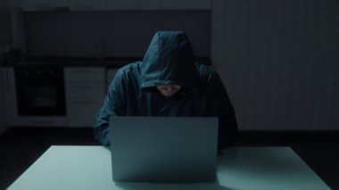 Kapüşonlu sweatshirt 'lü tanımlanamayan bir hacker, mutfakta dizüstü bilgisayarla çalışıyor. Tehlikeli ve ciddi bir hack sahnesinin tasviri..