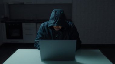 Mutfakta dizüstü bilgisayarla çalışan kapüşonlu bir adam. Siber güvenlik risklerini vurgulayarak tek başına çalışan bir hacker 'ın canlı bir temsili.