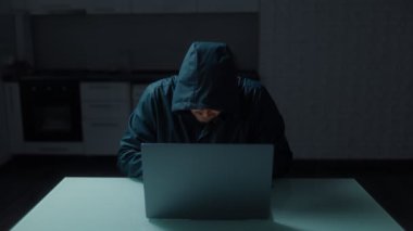 Karanlık bir odada bilgisayar korsanı, dizüstü bilgisayara odaklanmış. Görüntü, evde hacklemenin tehlikelerini vurgulayan bir aciliyet ve gizem hissi gösteriyor..