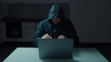Evdeki bir dizüstü bilgisayara odaklanmış bir adamın ciddi ve yoğun atmosferi. Kapüşonlu giyilen bu resim, hackerların gizli dünyasını temsil ediyor..