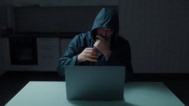 Kapüşonlu adam mutfakta oturuyor, dizüstü bilgisayara odaklanmış. Potansiyel olarak riskli bir siber güvenlik ortamında bir kişinin tasviri..