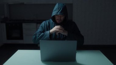 Kapüşonlu bir adam mutfak masasında tek başına oturuyor, dizüstü bilgisayarıyla meşgul. Görüntü, bilgisayar korsanlığı ile ilgili bir odak noktasını betimliyor.