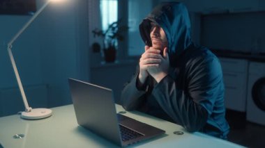 Kapüşonlu adam evde mutfaktaki dizüstü bilgisayara hızla yazmaya odaklanmış. Sahne karanlıkta çalışan gizemli bir hacker tasvir ediyor, siber risk ortamını vurguluyor..