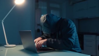 Loş bir mutfakta, bir adam bir masaya oturur, hızlı bir şekilde dizüstü bilgisayarda yazar. Kapüşonlu bir hacker 'ın bu yoğun anı bir ev ortamındaki tehlike ve aciliyeti yansıtır..