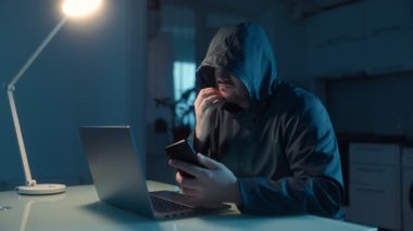 Kapüşonlu bir adam loş bir mutfakta, bir dizüstü bilgisayarın yanında oturuyor. Bu görüntü, tehlikeli bir hacker 'ın iş başındaki yoğun anını yakalıyor ve aciliyeti vurguluyor..