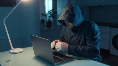 Kapüşonlu bir adam karanlık bir odada dizüstü bilgisayarla oturmuş, uzaktan kumandalı dolandırıcılıkla meşgul, dijital dünyadaki siber güvenlik ihlallerinin risklerini vurguluyor..