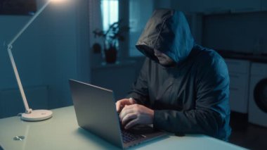 Kapüşonlu bir adam elinde bir dizüstü bilgisayarla masasında oturuyor, siber dünyadaki çevrimiçi güvenliğin modern zorluklarını vurguluyor..