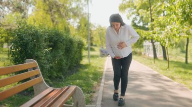 Hamile kadın, güneşli bir günde, yeşil bir parkta, karnı ağrıyan bir bankta oturmaya hazırlanırken, rahatsızlıkla karnını tutar..