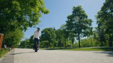 Kolları açık ve gülümseyen genç bir adam, parktaki ağaçlarla kaplı parlak bir patikadan aşağı doğru elektrikli bir tek tekerlekli bisiklette süzülüyor. Hareket ve özgürlük bir anda.