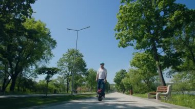 Kotlu ve miğferli bir adam, uzun ağaçlar ve sabah ışığı ile çerçevelenmiş, sakin bir parkta tek tekerlekli bisikletle sorunsuzca dolaşır..