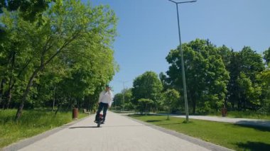 Genç adam sakin bir şekilde sakin bir parkta tek tekerlekli elektrikli bir araçta oturuyor. Kaskı ve kotu gündelik giyinmiş, ağaçlarla çevrili ve parlak güneş ışığı altında..