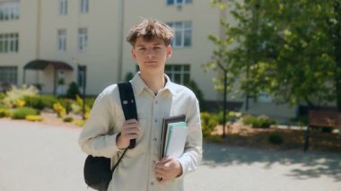 Lise öğrencisi, sıcak bir günde okul binasının önünde duruyor. Dosyalarını tutarken sakin ve kendinden emin görünüyor ve günlük giysiler giyiyor..