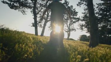 Bir adamın bacakları ve sırtı, çam ormanındaki uzun otların arasında elektrikli tek tekerlekli bisiklete binerken gösterilir. Yolculuk pürüzsüz, dengeli ve eğlenceli..