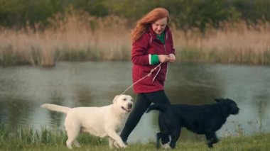 Kırmızı paltolu bir kadın, sakin bir göletin yanında duruyor. Biri siyah diğeri beyaz, iki neşeli köpekle takılıyor. Huzurlu doğanın tadını çıkarıyor..